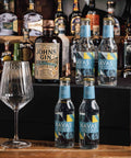 A bar-style product display featuring a bottle of Johnnie Walker Pink Label Gin, two Navas sparkling waters bottles, and an empty wine glass arranged on and around a black box in front of a blurred liquor shelf.