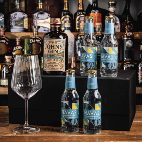 A bar-style product display featuring a bottle of Johnnie Walker Pink Label Gin, two Navas sparkling waters bottles, and an empty wine glass arranged on and around a black box in front of a blurred liquor shelf.