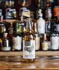 A bottle of Cornish Cock Craft Cider with a white label featuring a black rooster illustration, standing on a wooden bar counter with blurred liquor bottles in the background.