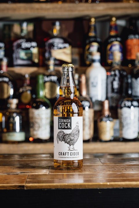A bottle of Cornish Cock Craft Cider with a white label featuring a black rooster illustration, standing on a wooden bar counter with blurred liquor bottles in the background.