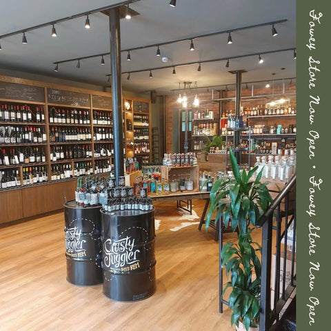 A liquor store interior with shelves of wine and spirits, two black barrel-style display cases labeled 'Custy Juggler' in the foreground, wood flooring, track lighting, and a green vertical banner with store text along the right edge.