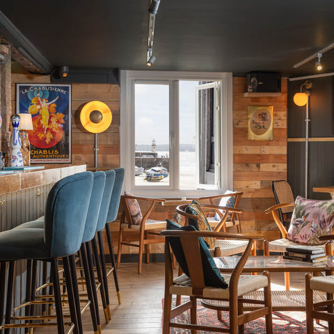 A cozy, rustic indoor lounge or bar setting with wood-paneled walls, a row of blue velvet bar stools, wooden dining chairs with floral cushions, and a large window looking out onto water and a lighthouse.