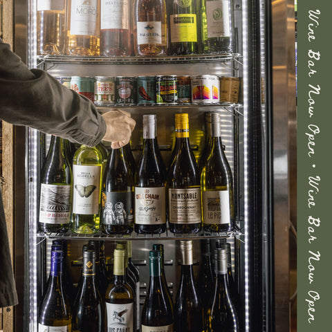 A person's arm reaches into a glass-front wine refrigerator stocked with assorted wine bottles and canned beers. The scene is lit by the refrigerator interior, with a vertical green sign on the right reading 'Wine Bar Now Open'.