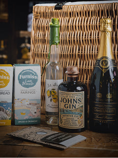 A curated gift basket arrangement on a wooden surface featuring a large woven wicker basket, a bottle of Johnnie Walker Black Label Gin, a bottle of Camembert de Camberley Valley, a box of Furniss Blue Mountains Fairings, a loaf of bread, and a bottle of tea, with a gift bag in the foreground.