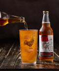 A bottle of Cornish Scrumpti craft cider and a matching pint glass with a rooster graphic, with amber liquid being poured into the glass on a dark rustic wooden surface.