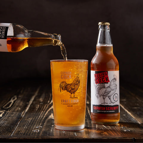 A bottle of Cornish Scrumpti craft cider and a matching pint glass with a rooster graphic, with amber liquid being poured into the glass on a dark rustic wooden surface.