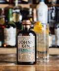 A bottle of St. Ives John's Gin stands on a wooden bar counter next to a tall glass filled with a clear cocktail and a lemon twist, with blurred bottles in the background.