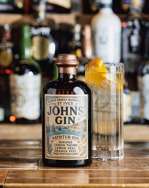 A bottle of St. Ives John's Gin stands on a wooden bar counter next to a tall glass filled with a clear cocktail and a lemon twist, with blurred bottles in the background.
