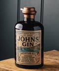 A dark glass bottle of St Ives Johns Gin with a vintage-style label, resting on a rustic wooden surface against a dark paneled background.