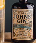 A bottle of St Ives John's Gin Bath Tub Gin with a vintage-style cream label, shown on a wooden surface with a blurred glass of gin and lemon slice in the foreground.