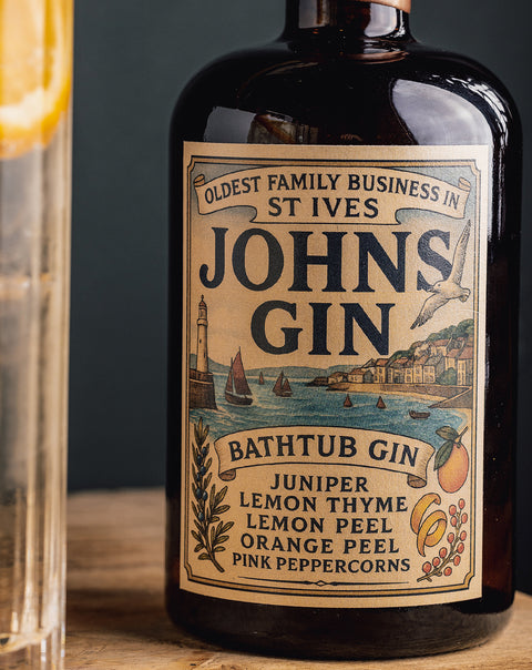 A bottle of St Ives John's Gin Bath Tub Gin with a vintage-style cream label, shown on a wooden surface with a blurred glass of gin and lemon slice in the foreground.