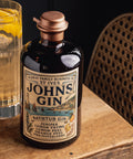 A bottle of Johns Gin Bathtub Gin with a copper-toned cap and vintage-style label, placed on a wooden surface next to a glass of amber liquid, with a woven chair in the background.