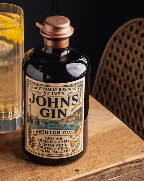 A bottle of Johns Gin Bathtub Gin with a copper-toned cap and vintage-style label, placed on a wooden surface next to a glass of amber liquid, with a woven chair in the background.