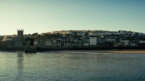 A scenic coastal town view with a church tower on the left, a hillside village with white buildings, and calm water in the foreground under a clear blue sky.
